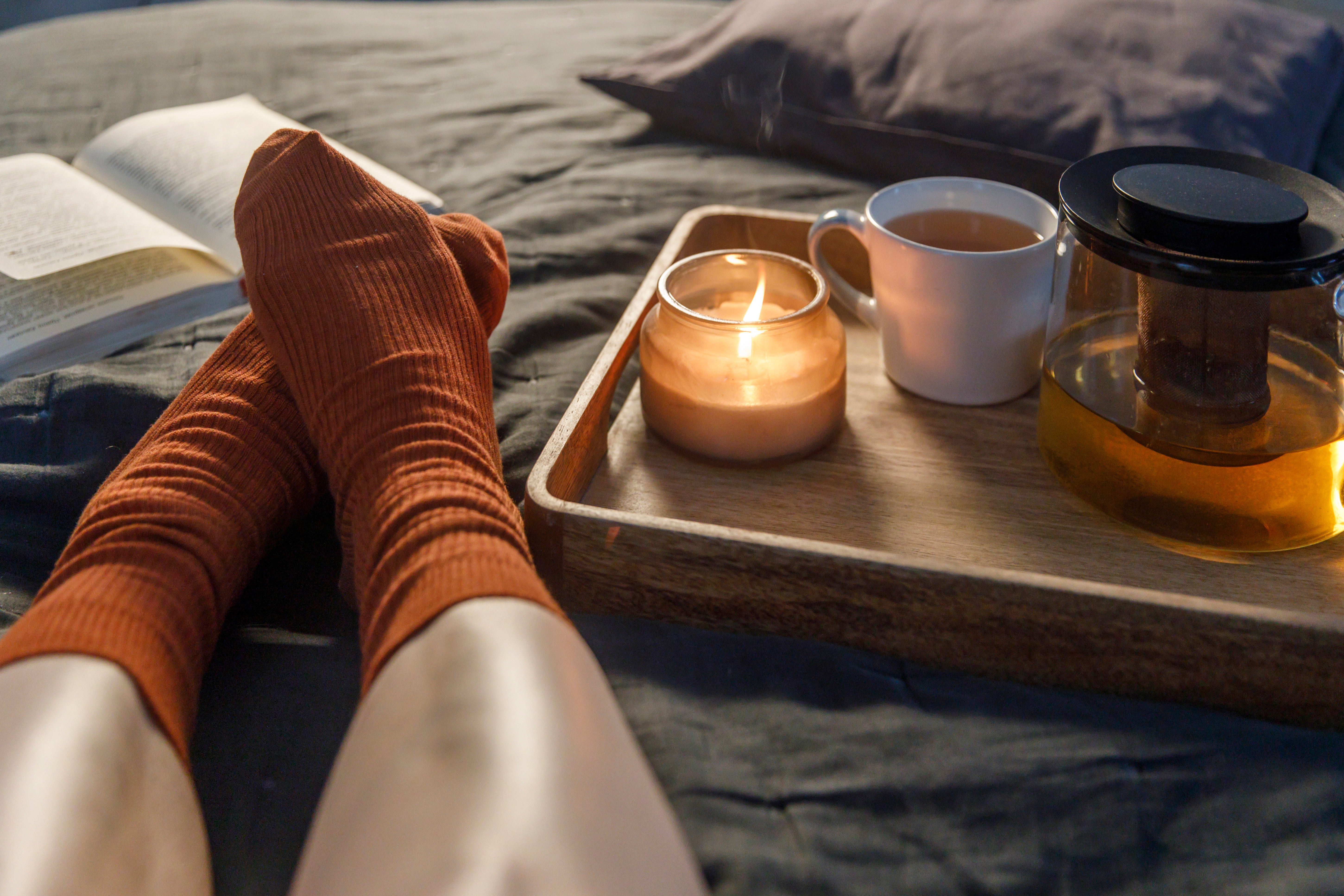 Legs of a woman laying on the bed wearing orange socks. Besides the feet is a tablet with a candle and a cup and pot of tea.